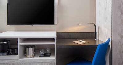 Detailed View of TV Above Hospitality Center, Work Desk With Illuminated Lamp, and Blue Task Chair in Guest Room