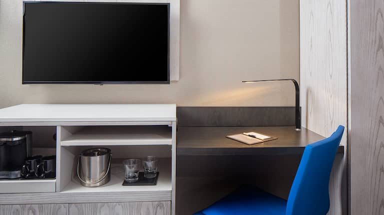 Detailed View of TV Above Hospitality Center, Work Desk With Illuminated Lamp, and Blue Task Chair in Guest Room