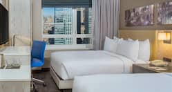 Wall Art Above Two Queen Beds, Illuminated Lamp, Bedside Table, Hospitality Center, TV, Work Desk With Blue Chair, and Window With Open Drapes to City View in Guest Room