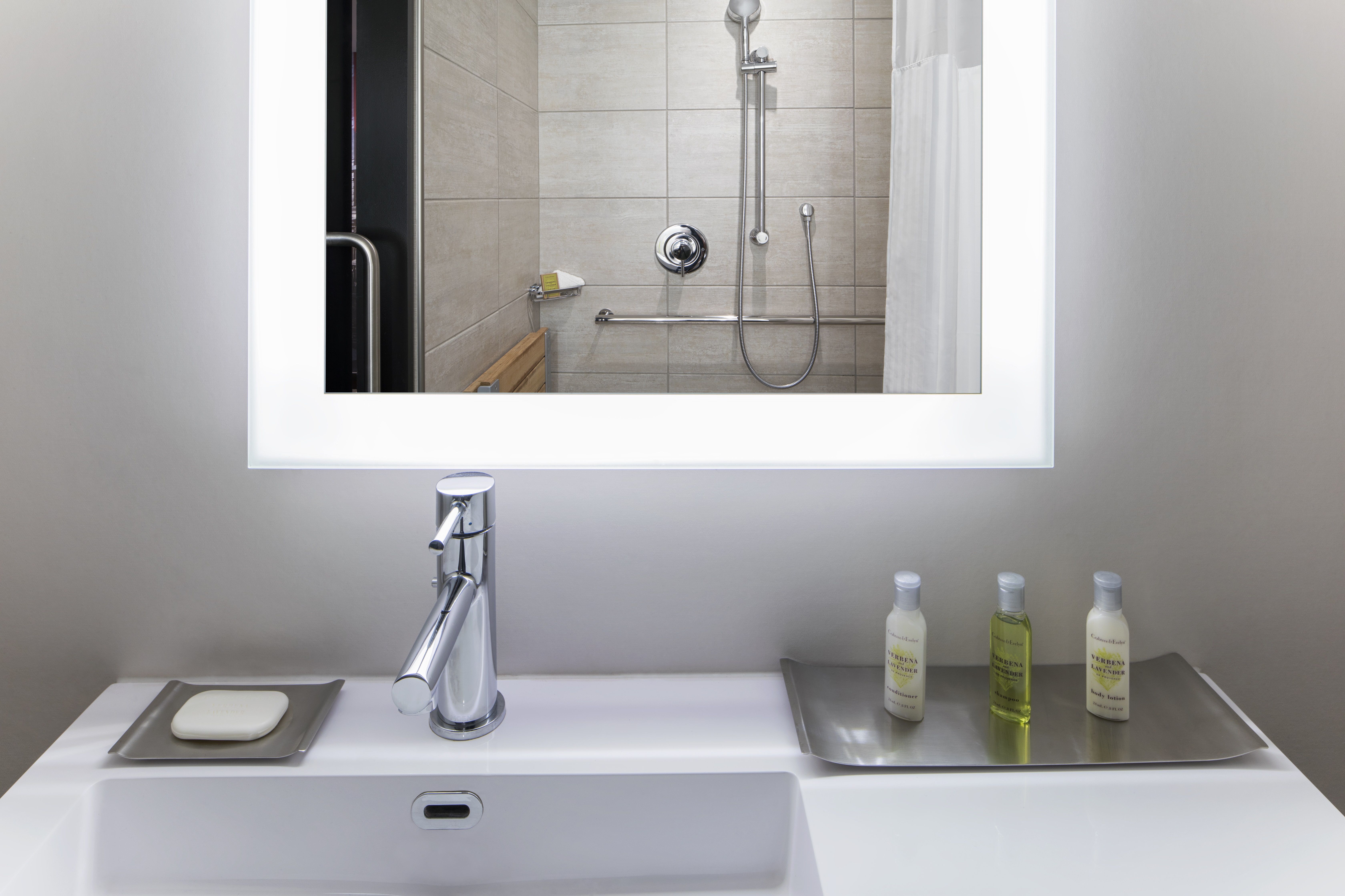 Shower With Seat, Grab Bars and Handheld Showerhead Reflected in Brightly Lit Vanity Mirror, Sink, and Toiletries in Accessible Bathroom