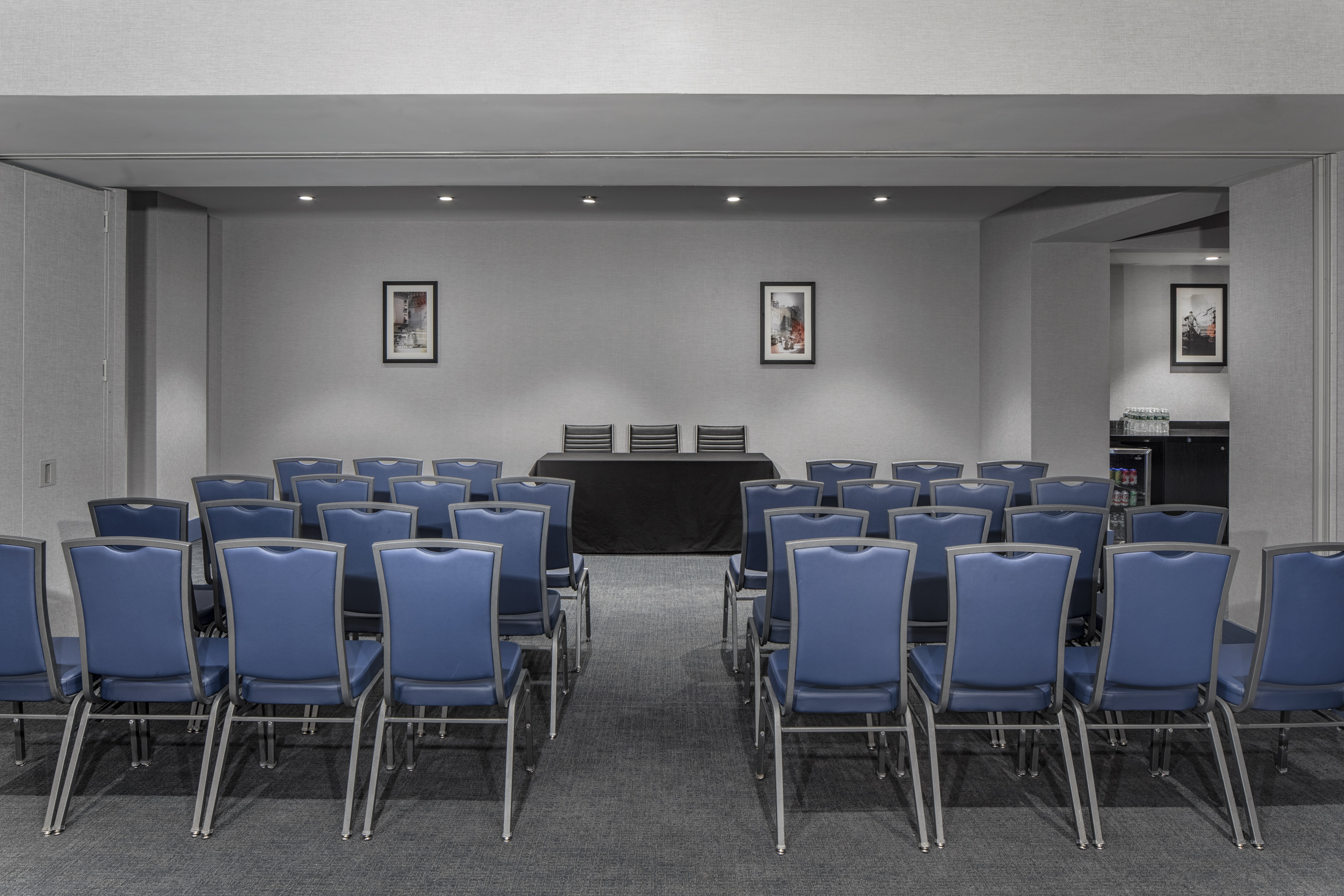 Meeting Room Arranged Theater Style With Rows of Blue Chairs Facing Wall Art ans Speaker's Table With Two Chairs and View of Refreshment Area