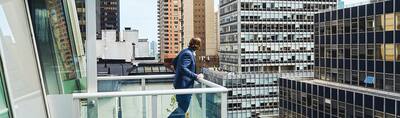 A man in a suit stands on a balcony looking out to a built up city view