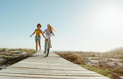 Two women on a boardwalk near the beach, one riding a bike, the other running towards the ocean