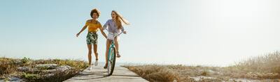 Two women on a boardwalk near the beach, one riding a bike, the other running towards the ocean