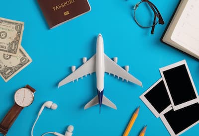 A blue background set against various holiday-related objects including a model airplane, a passport, money, Polaroid photos, and a watch. 