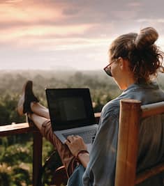 Woman on a laptop on a balcony looking out to a forest.