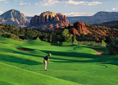 A golfer taking a swing on a bright green gold course with red mountains in the background.