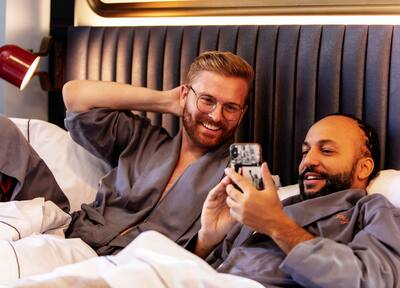 Two men in bed laughing and smiling at something on the cell phone