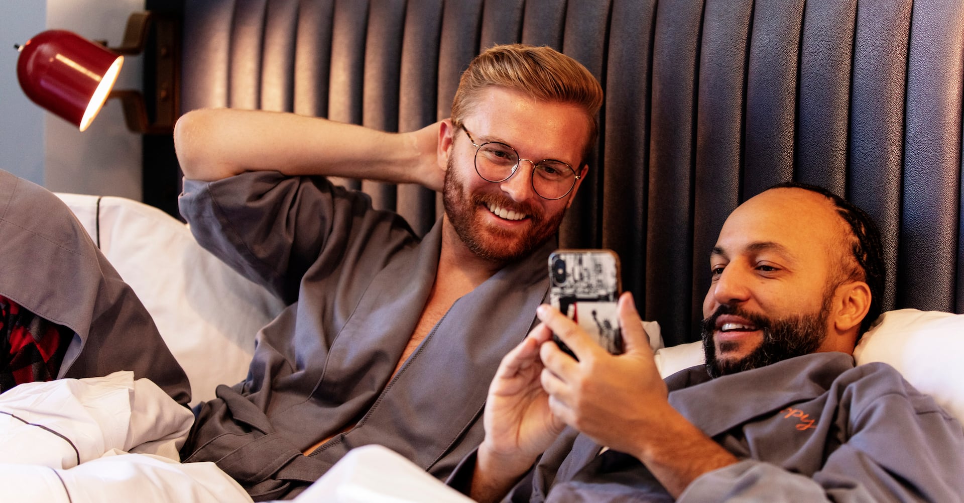 Two men in bed laughing and smiling at something on the cell phone