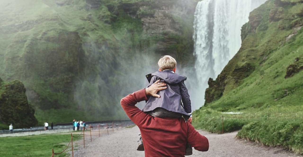 Family on vacation in Iceland. Father carries a small son on his shoulders to the famous waterfalls of Skogafoss.