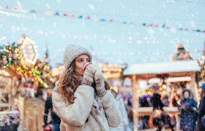 Girl drinking hot coffee while walking in Christmas market decorated with holiday lights in the evening.