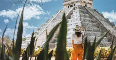 A young woman tourist in a hat stands against the background of the pyramid of Kukulcan in the ancient Mexican city of Chichen Itza. 