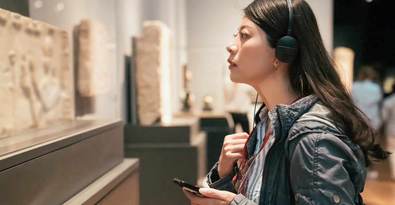 curious young asian chinese woman tourist looking at exposition in museum using headphones and listening audio guide in modern gallery.