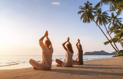 Group of three people practicing yoga lotus position on the beach for relaxation and wellbeing, warm tropical summer landscape with palm trees