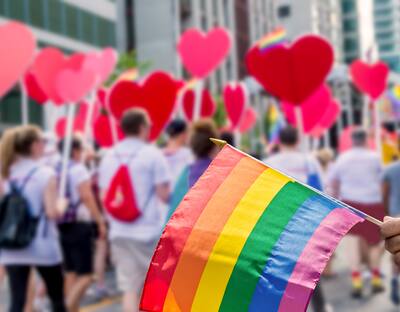 Hand holds a pride flag in front of a street of people with heart shaped signs