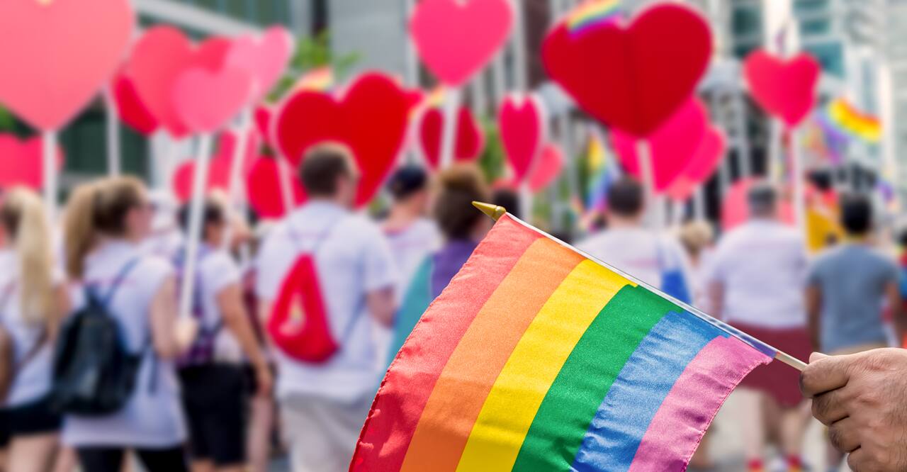 Hand holds a pride flag in front of a street of people with heart shaped signs