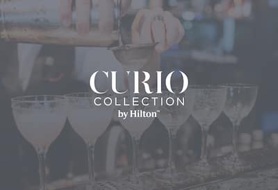 A bartender pours cocktails into 6 glasses lined up on a bar. The Curio Collection by Hilton logo is layered on top of the image