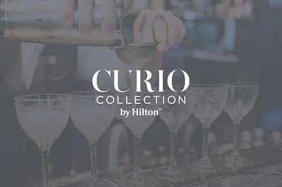 A bartender pours cocktails into 6 glasses lined up on a bar. The Curio Collection by Hilton logo is layered on top of the image