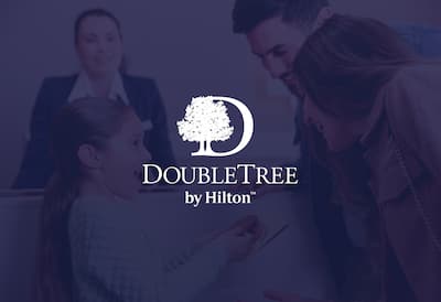 A young girl and her parents receiving a DoubleTree cookie at check in. The DoubleTree by Hilton logo is layered over the image