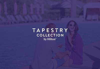 A laughing couple share a drink by the pool. The Tapestry Collection by Hilton logo is layered over the top of the image