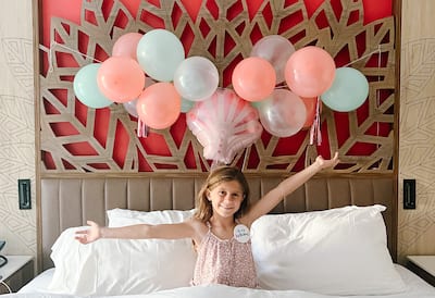 Girl sits in bed with arms outstretched with balloons above her head