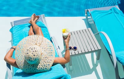 Woman sits next to pool in hat with drink