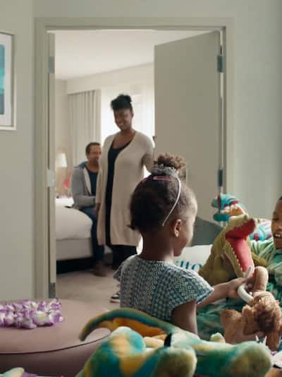 A family in a hotel room with the parents coming out of the bedroom and the children sitting in the living room
