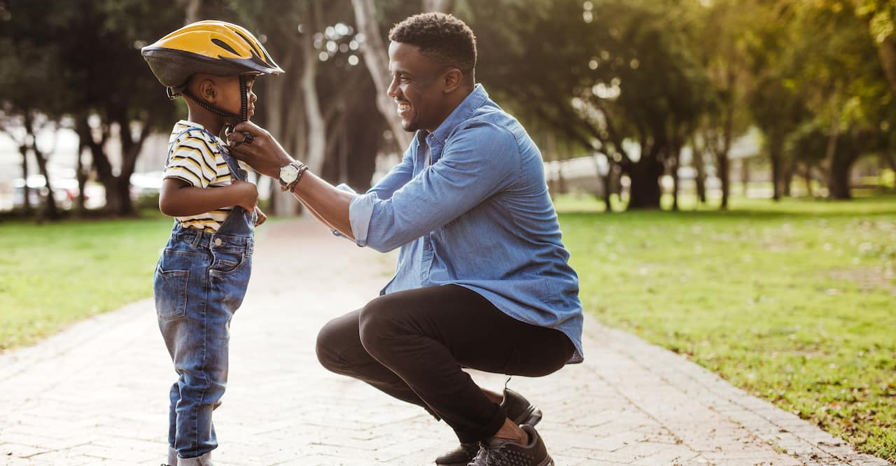 African man putting helmet on cute boy at the park. 