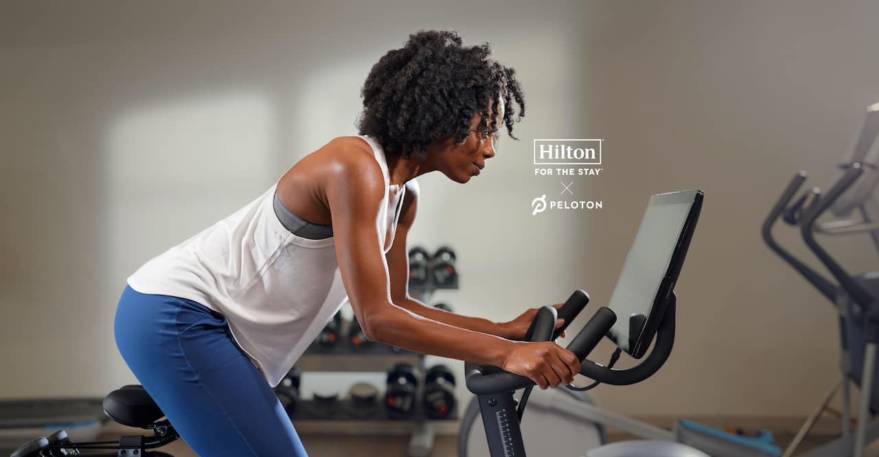 A woman on Peloton bike in a Hilton fitness center. Hilton and Peloton logos are visible in the middle of the image.