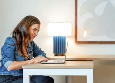 Woman working on laptop while sat at pull out desk within guest room.
