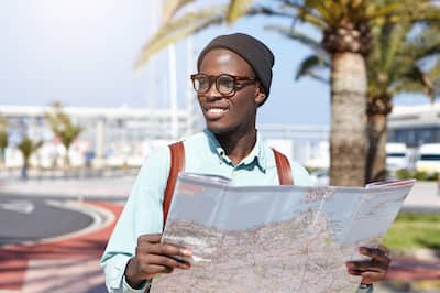An African American man wearing glasses, a black cap, a blue button down shirt and a backpack reviewing a map while standing in front of palm trees.