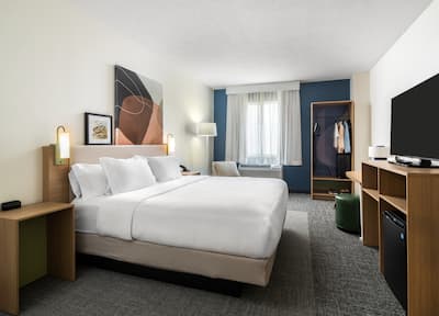 A spacious hotel room featuring a comfortable bed in the center. The room features a modern design with a grey carpet, blue feature wall, light grey curtains, an open closet, wood table tops, plus orange and blue artwork.