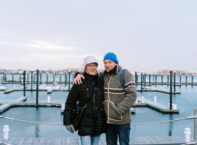 Couple in winter jackets and hats smiling in front of the Boston Harbor.