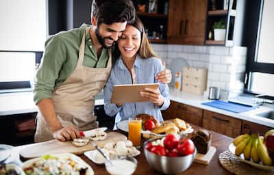 Couple stands in kitchen with food looking at tablet 