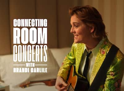 'Connecting Rooms Concerts with Brandi Carlile' text overlay on an image of Brandi Carlile sitting on a bed playing acoustic guitar