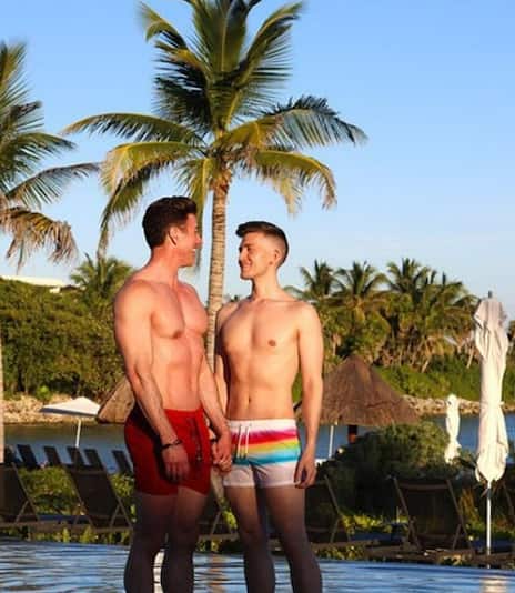 A gay male couple holding hands while standing in the pool. Palm trees and clear skies are in the background.