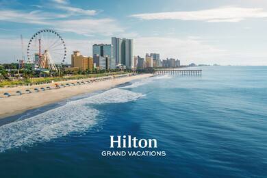 View of ocean with a pier, buildings, and a ferris wheel. Hilton Grand Vacations logo is overlaid.