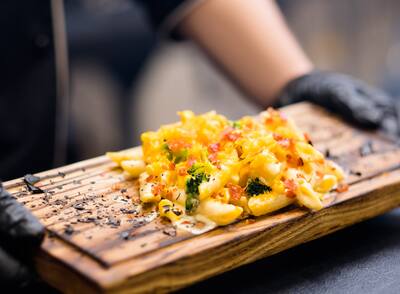Chef holding mac and cheese with broccoli and bacon bits on wooden board.
