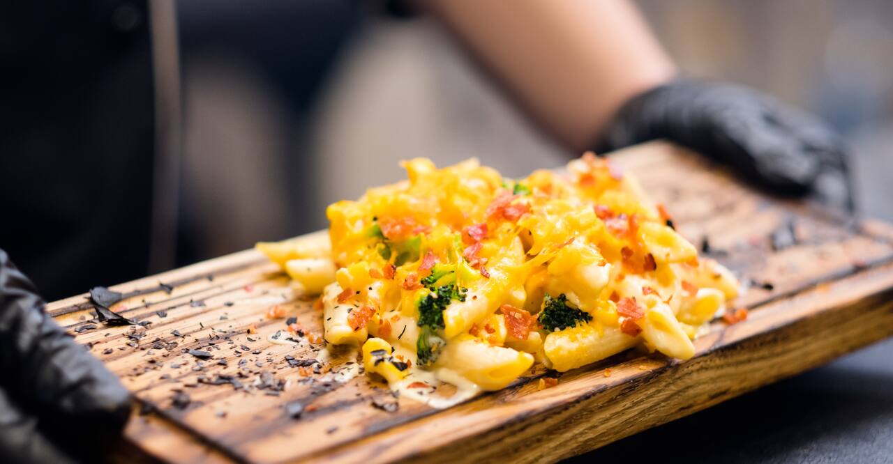 Chef holding mac and cheese with broccoli and bacon bits on wooden board.