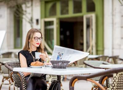 Young woman having a breakfast with coffee and croissant reading newspaper outdoors at the typical french cafe terrace in France