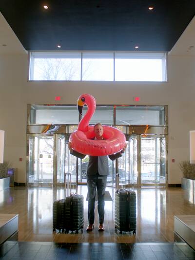 A man with a flamingo pool raft over his head in a business suit in the lobby of a hotel.