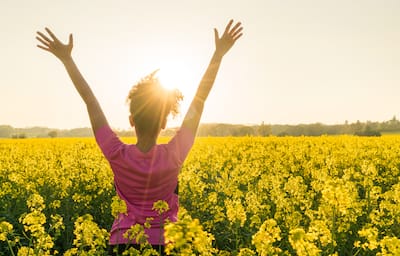 Young girl raising her arms in the sky as she stands in the middle of a flowery meadow during sunset.
