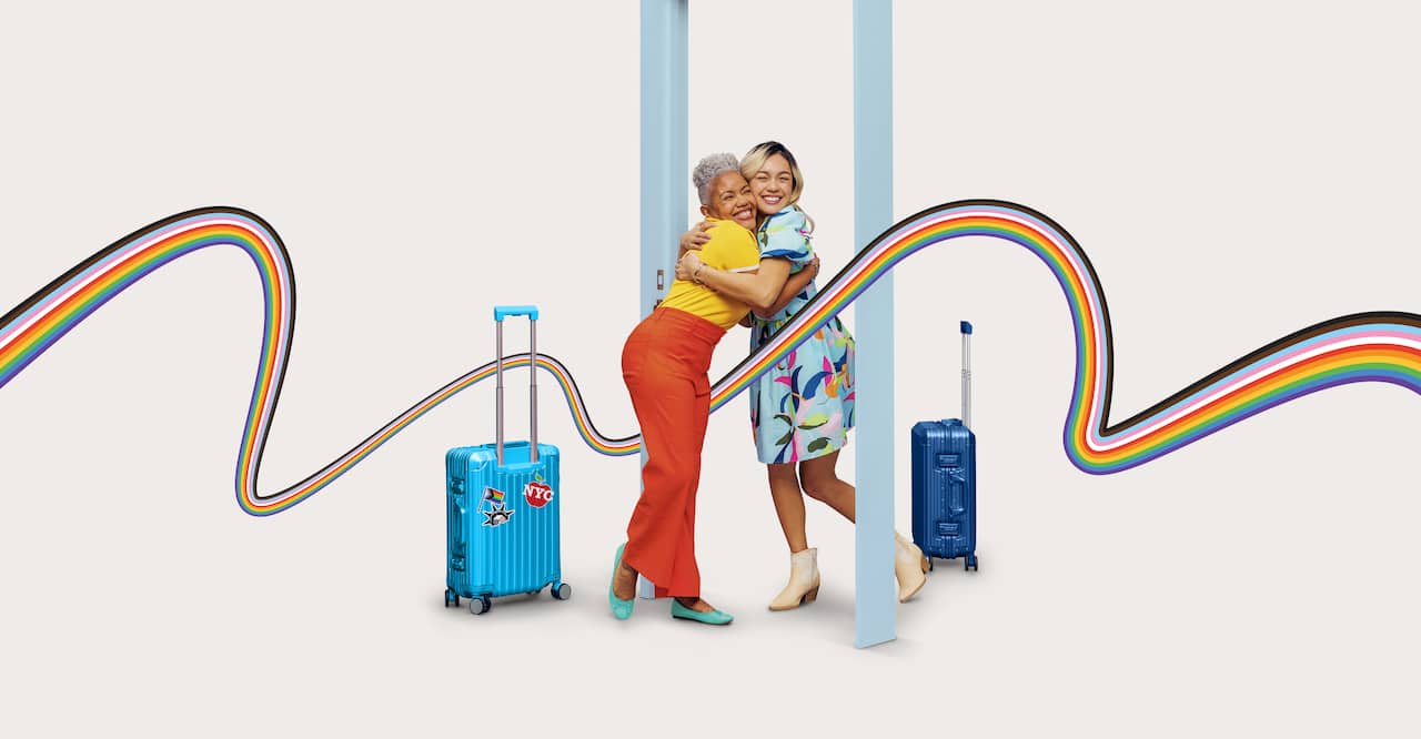 An LGBTQ+ couple embracing with smiles on their faces. Both of them are standing underneath a doorframe with their suitcases at either side of them. A rainbow ribbon graphic is scattered across the screen.