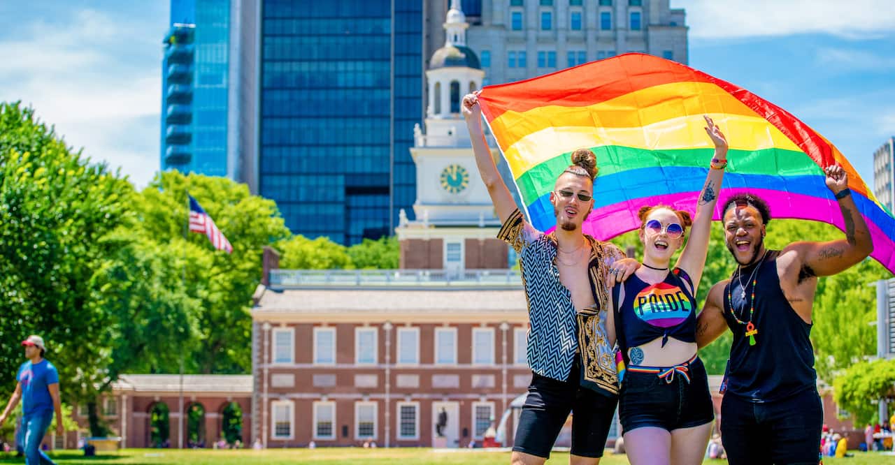Three LGBTQ supporters holding a rainbow flag in front of Independence Hall in Philadelphia.