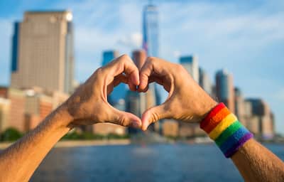 Hands wearing gay pride rainbow sweat band making heart symbol in front of city skyline.