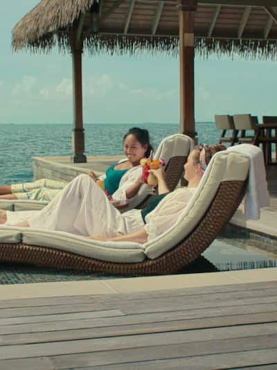 Two woman on sun loungers on an oceanside deck with thatched roof smiling and drinking cocktails