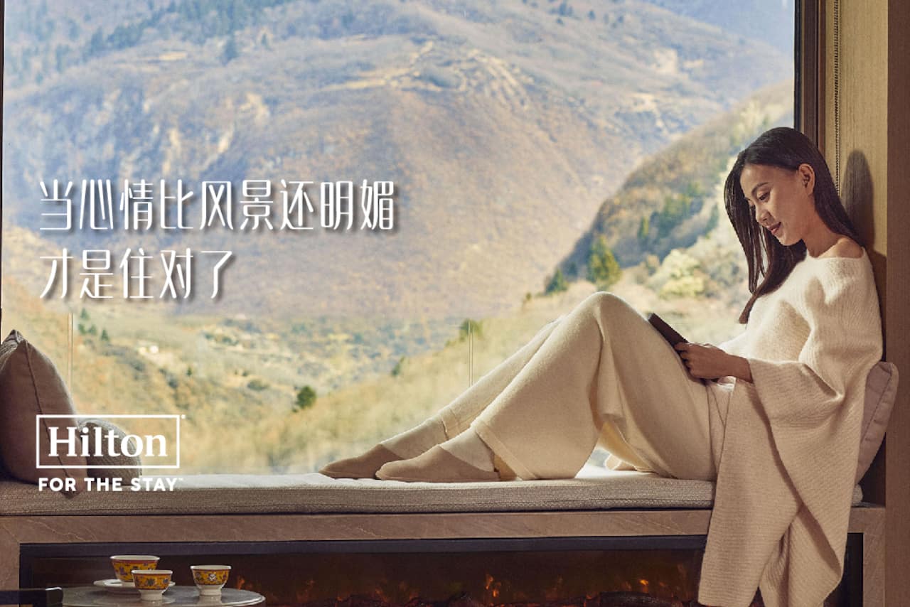 Woman sitting on a window ledge reading a book. Hilton For The Stay is stated in the bottom left hand corner.