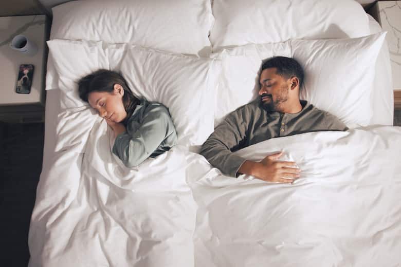 A couple rests peacefully in a hotel bed with crisp white linens. On the nightstand, a coffee mug and a phone displaying a dog photo add a personal, cozy touch.