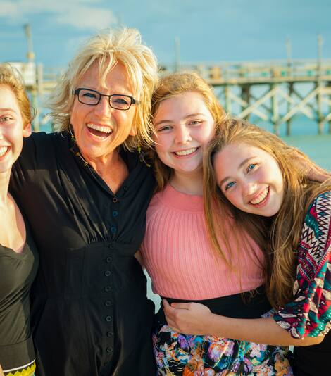 Three young girls hug an older woman on a beach in front of a pier. 