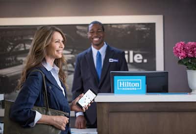 Woman holding cell phone showing room options with front desk reception team member in background